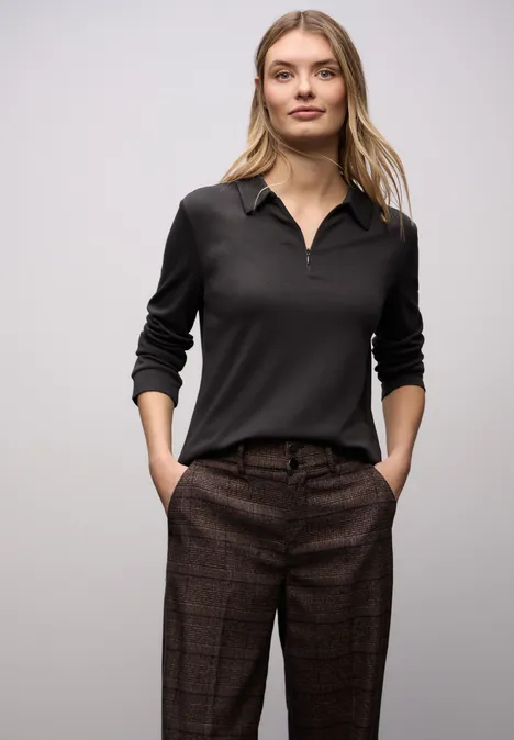 Frau in schwarzem Langarm-Poloshirt und karierter brauner Hose, Hände in den Taschen.